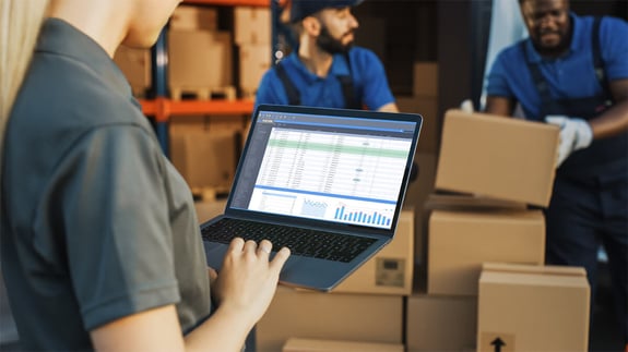 Woman using software to track logistics KPIs and two men moving boxes